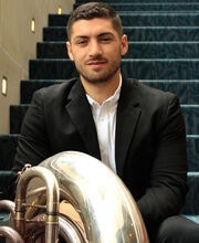 Jordan Oliveira sitting on a carpeted stairway with his tuba. He has short dark brown hair, mustache and beard and is wearing a black suit over a white button down shirt