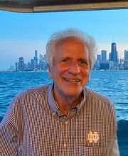 Leonard Paolillo on a boat smiling with short gray hair wearing a black and white checked button down shirt with water and a city skyline in the background