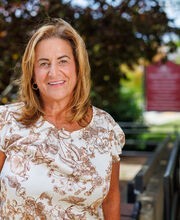 Kathleen Perry standing in front of a bench on campus smiling with medium length brown hair with blonde highlights and wearing an off-white with brown floral print short sleeve top