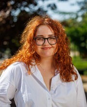 Halle Singh smiling with long curly red hair and wearing large black rimmed glasses and a white long-sleeve button up shirt