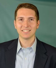 Philip Szlosek smiling with short brown hair and wearing a navy blue blazer over a blue and white pinstripe button up shirt