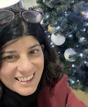 Sarah Thomas smiling with shoulder length wavy dark brown hair wearing a pair of purple rimmed glasses on top of her head and a red blazer over a black top