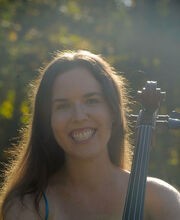 Dr. Sara Wilkins smiling with sun shining on her long dark brown hair. She is wearing a blue spaghetti strap top and the neck of her cello is in front of her shoulder
