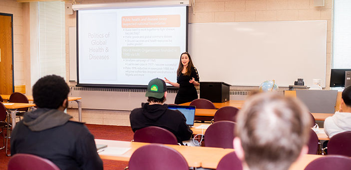 Professor Sidita Kushi teaching at front of classroom in front of a projector screen while students look on
