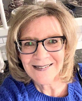 Donna Abelli smiling with medium length dirty blonde hair and wearing black rim glasses and a royal blue sweater