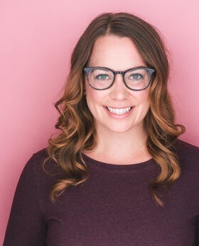 Sarah Bedard smiling with long wavy light brown hair and wearing large rimmed glasses and a long sleeve plum colored jersey