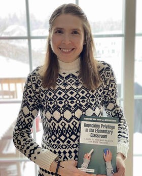 Dr. Jacquelynne Boivin smiling with long light brown hair and wearing a black and white sweater. She is holding her book titled Unpacking Privilege in the Elementary Classroom