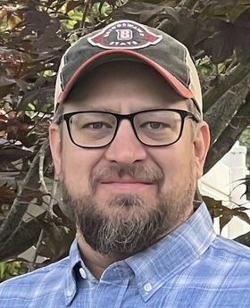 Emmett Buhmann with short gray brown hair, mustache and beard wearing black rim glasses a green and khaki baseball cap and a blue and white plaid button down shirt