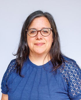 Dr. Susan Flis smiling with medium length straight graying brown hair and wearing brown rimmed cat's eye shaped glasses and a short sleeve blue top