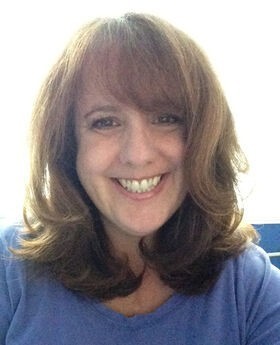 Sheila J. Gibbons smiling with long light brown wavy hair wearing a blue crew neck top