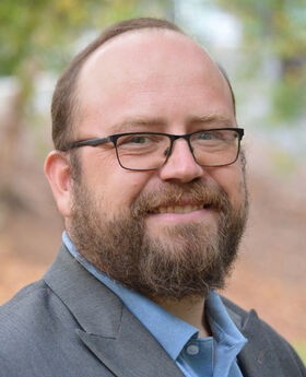 Dr. Feodor Gostjev smiling with short balding reddish brown hair, mustache and beard, wearing black rim glasses and a gray blazer over a light blue button up shirt