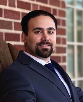 Dr. Kianoosh Hassani smiling with short dark brown hair, mustache and beard and wearing a navy blue suit jacket and tie with button down white shirt