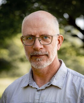 John Kucich balding with short gray hair, mustache and beard and wearing a light blue button up shirt with 2 front pockets