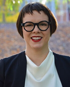 Dr. Melanie McNaughton with short dark brown hair wearing black rim round glasses, a white blouse and black jacket