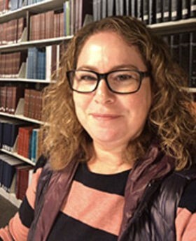 headshot photo of Linda Meiberg in front of library bookshelves with light brown medium length curly hair wearing black rim glasses and a black and salmon top under a black vest