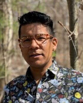 headshot photo of Dr. Luis Paredes with short wavy dark hair, wearing light brown glasses and a floral button down shirt with a dark background. Woods are