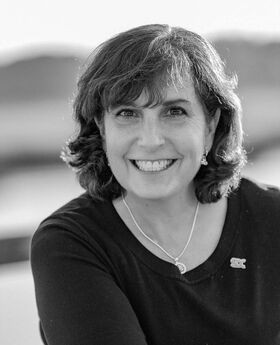 black and white photo of Lisa Rafferty smiling with shoulder wavy brown hair, leaning on her forearm and wearing a round neck dark colored three-quarter sleeve jersey
