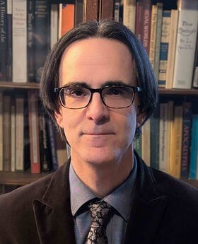 Dr. J.R. Webb with medium length salt and pepper straight hair wearing brown rim glasses, a blue button down shirt with a dark brown blazer and dark brown paisley tie
