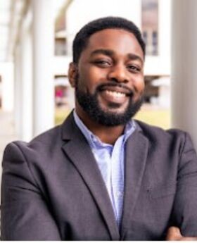 Dr. Maurice Williams Jr. smiling with short black hair, mustache and beard and wearing a light blue button down shirt under a gray suit jacket