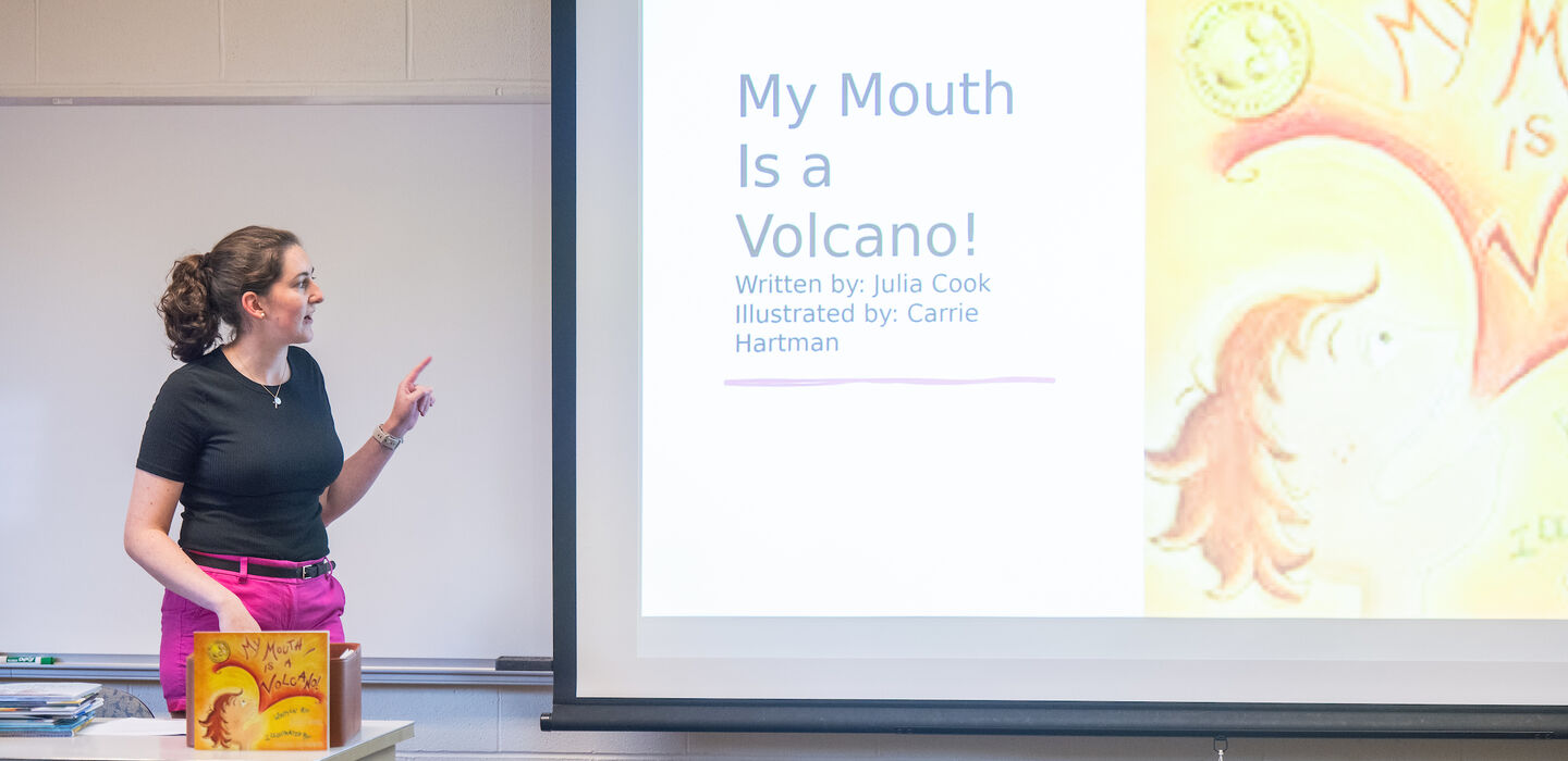 Elementary Education teacher in classroom pointing at projector screen with cover and info about a book that is displayed on the desk in front of her