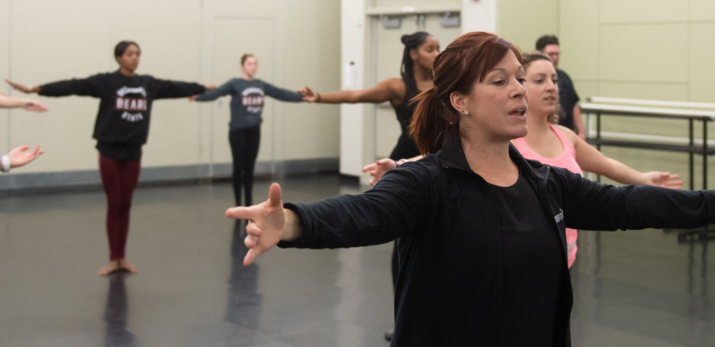 a ballet class with the professor in the foreground and students across the room behind her, all with their arms open wide and their legs together in a dance position
